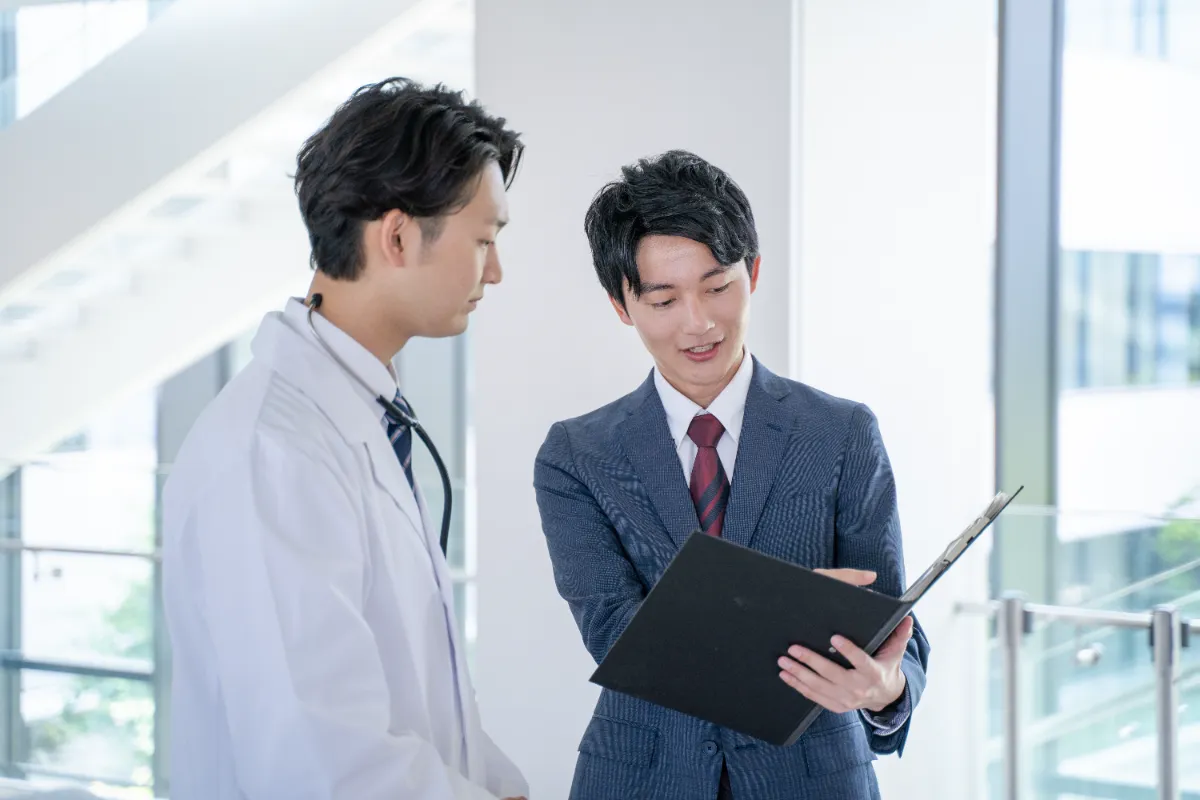 病院の廊下で書類を手に打ち合わせをする白衣の医師とビジネスパーソン